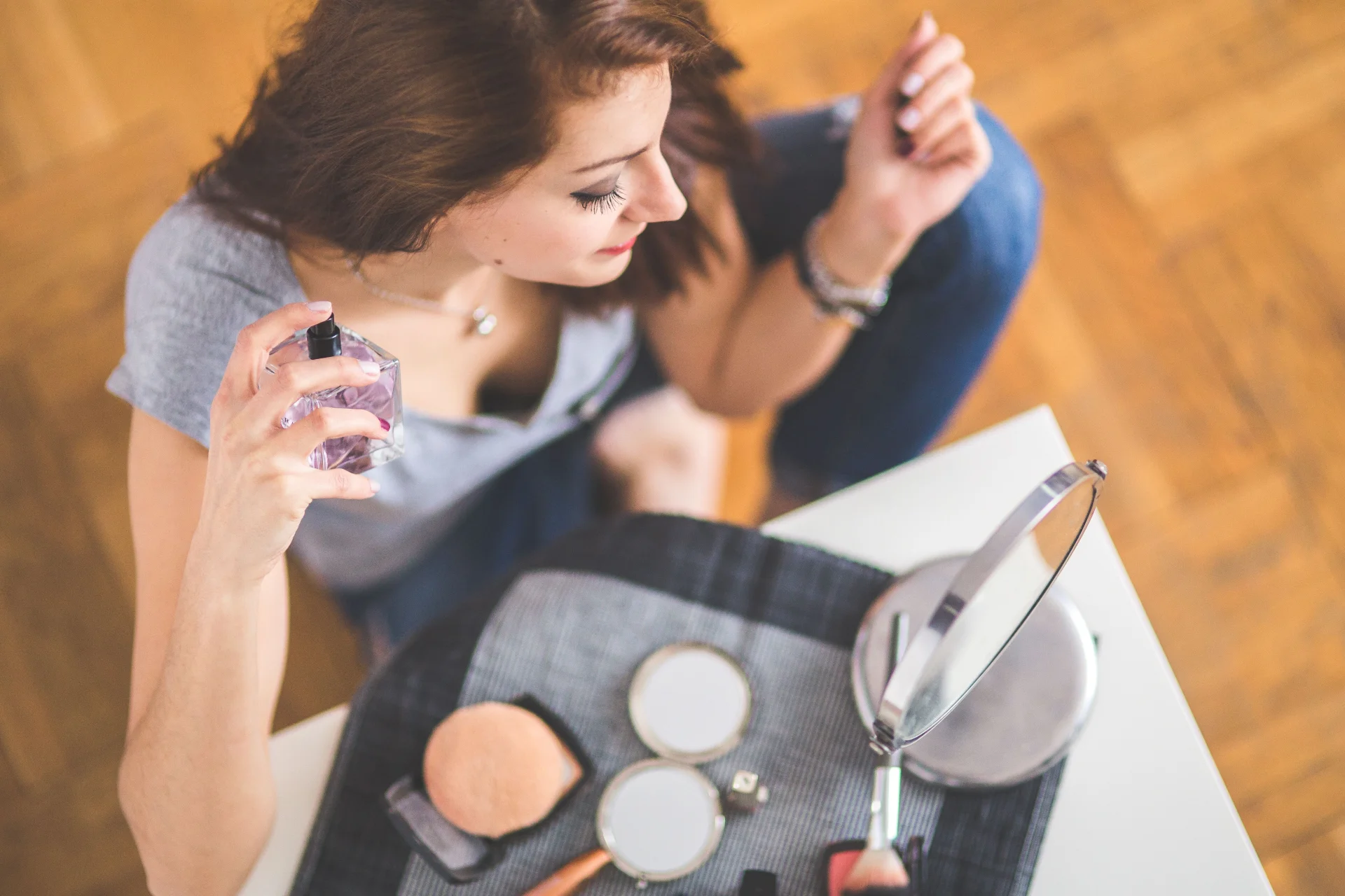 Młoda kobieta aplikuje perfumy, wokół niej leżą niezbędne kosmetyki do makijażu.